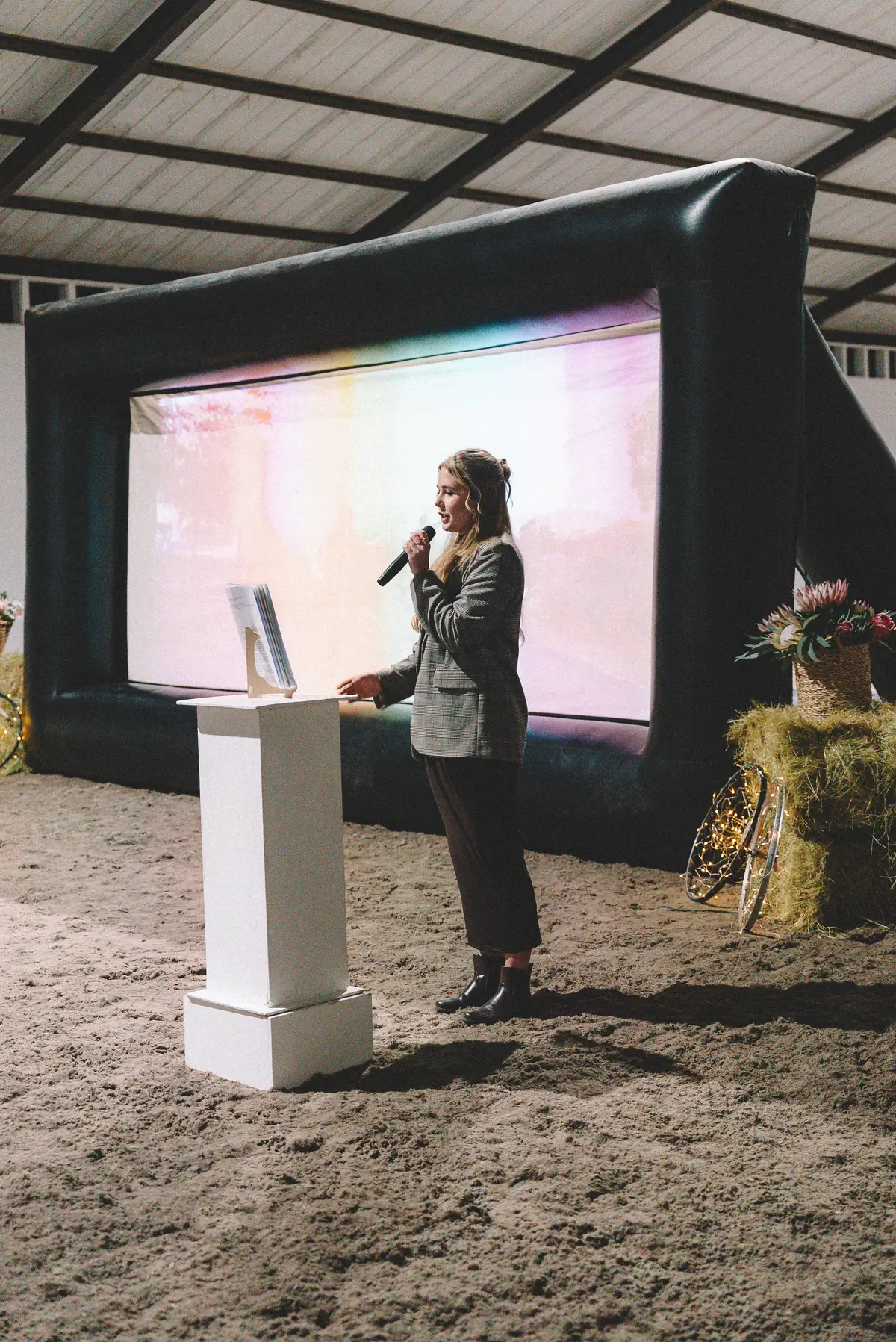 A host introducing the film beside the big screen in the indoor arena, hay bales and proteas either side