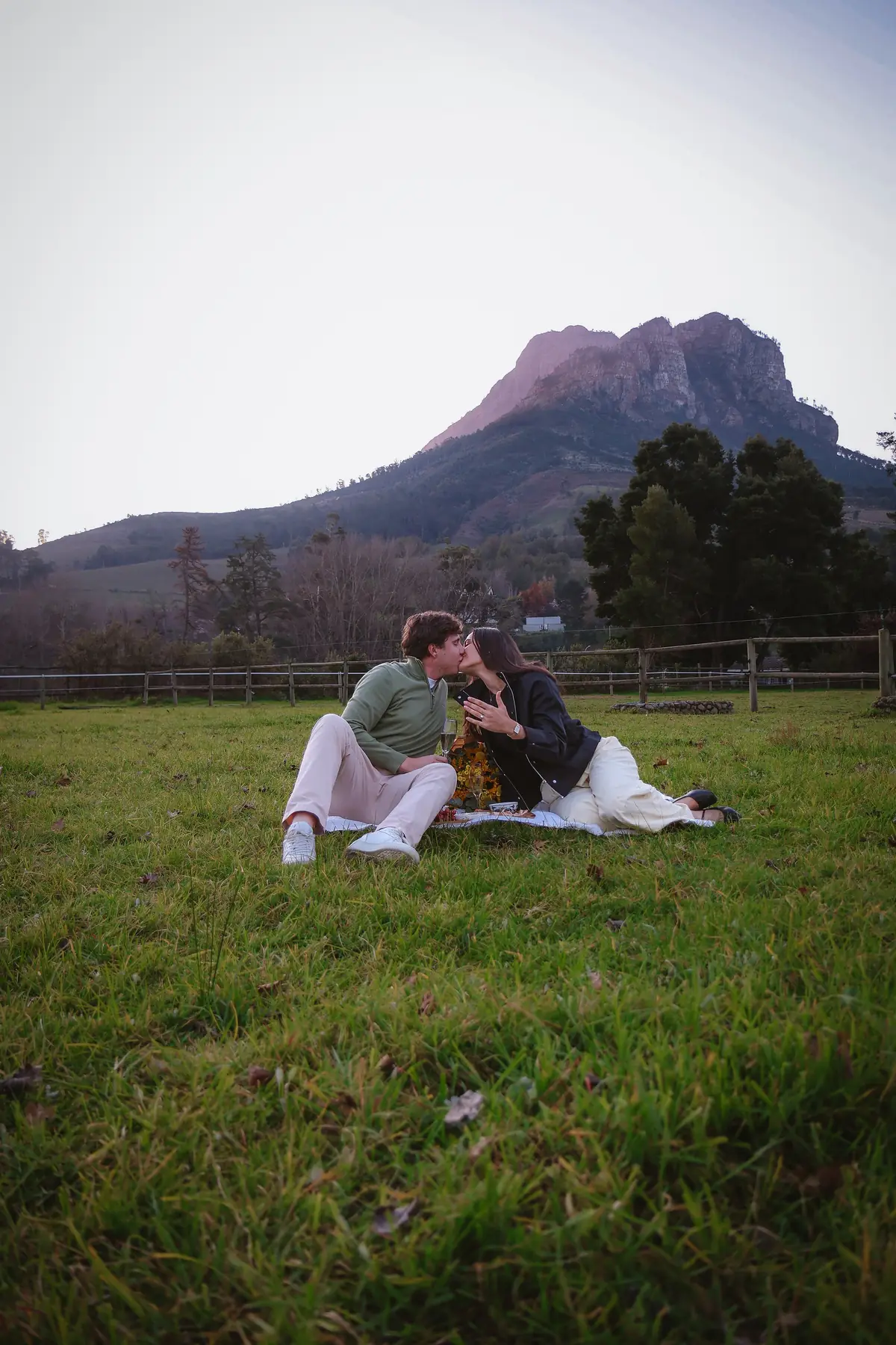 A newly engaged couple seated in the meadow at Banhoek Stud at golden hour, mountain peak behind them